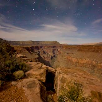 A moonlit scene of a cracked boulder hanging over the top of the Grand Canyon below.  